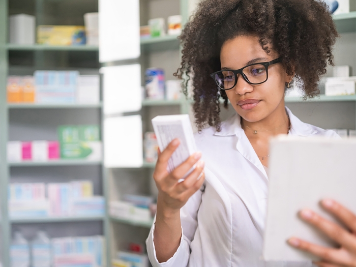 Person looking at a box of medication with a tablet in their hands standing in front of a shelf of medication.