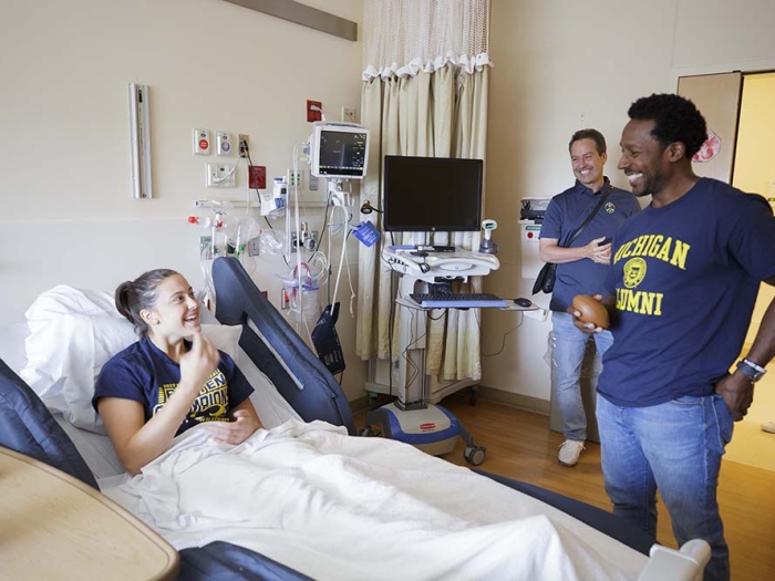 Desmond Howard visits a patient at Mott. He is smiling and wearing a U-M alumni t-shirt and jeans. The patient is lying on a hospital bed and is covered with a white blanket. 