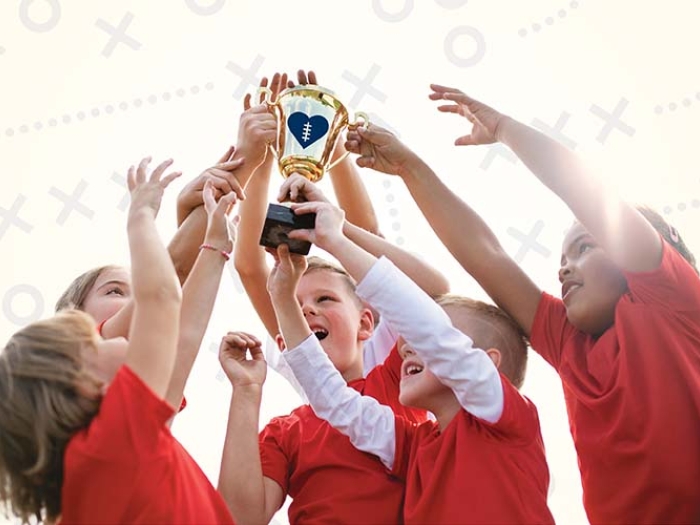 Five children in red t-shirts standing in a tight group with their arms extended upwards to hold up a gold trophy with a blue heart.