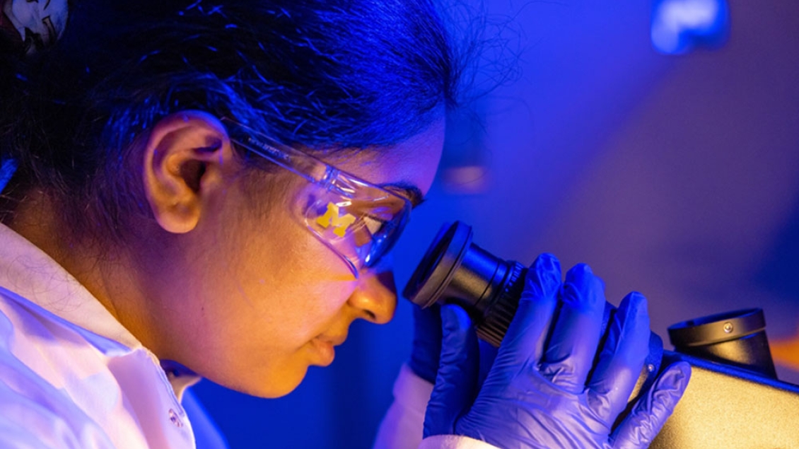 A woman wearing safety glasses looks into a microscope.