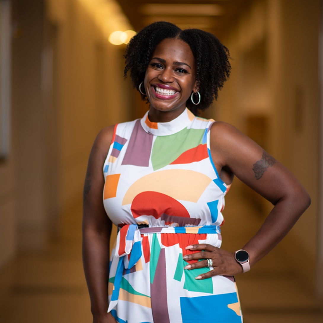 Black woman wearing dress with multi-colored abstract shapes and hoop earrings, smiling with one hand on hip