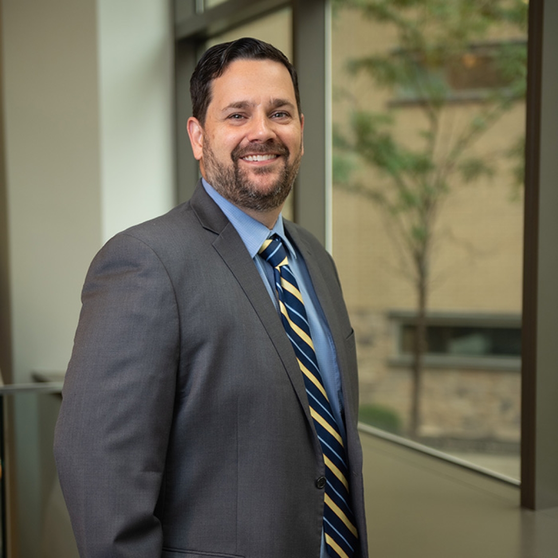 White male with brown hair and beard wearing suit jacket, blue shirt and striped tie